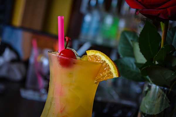 Orange cocktail garnished with cherry and orange slice, pink straw, red rose in background, warm colors, festive mood