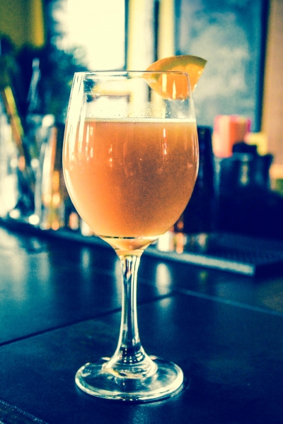 Wine glass with orange-hued cocktail garnished with a citrus wedge on a dark bar top, warm tones, relaxed mood