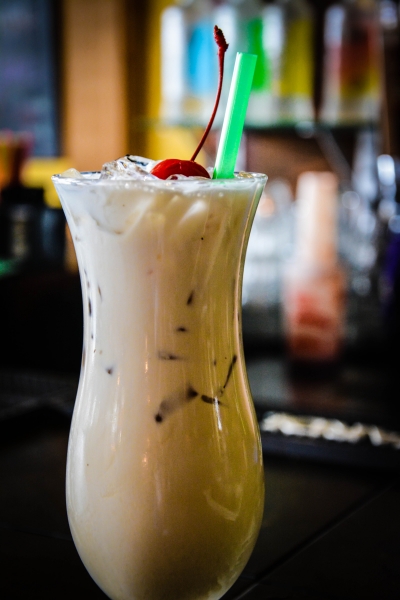 Creamy pale cocktail with cherry garnish and green straw on a dark bar, soft cream and dark tones, cozy mood