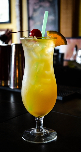 Tall hurricane glass cocktail with yellow-orange drink, garnished with cherry, orange slice, and green straw, on dark bar