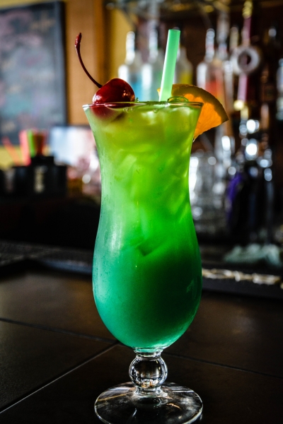 Bright green cocktail with cherry and orange garnish and straw, dark bar background with bottles, vivid green tones, festive mood