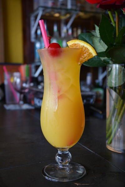 Orange tropical cocktail in a hurricane glass with cherry, orange slice, and pink straw, bar backdrop, red flowers in vase