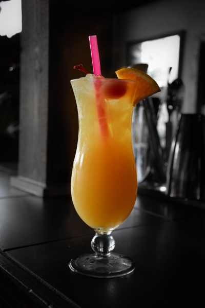 Orange tropical cocktail with cherry and orange garnish, pink straw, dramatic dark and warm lighting, bar setting