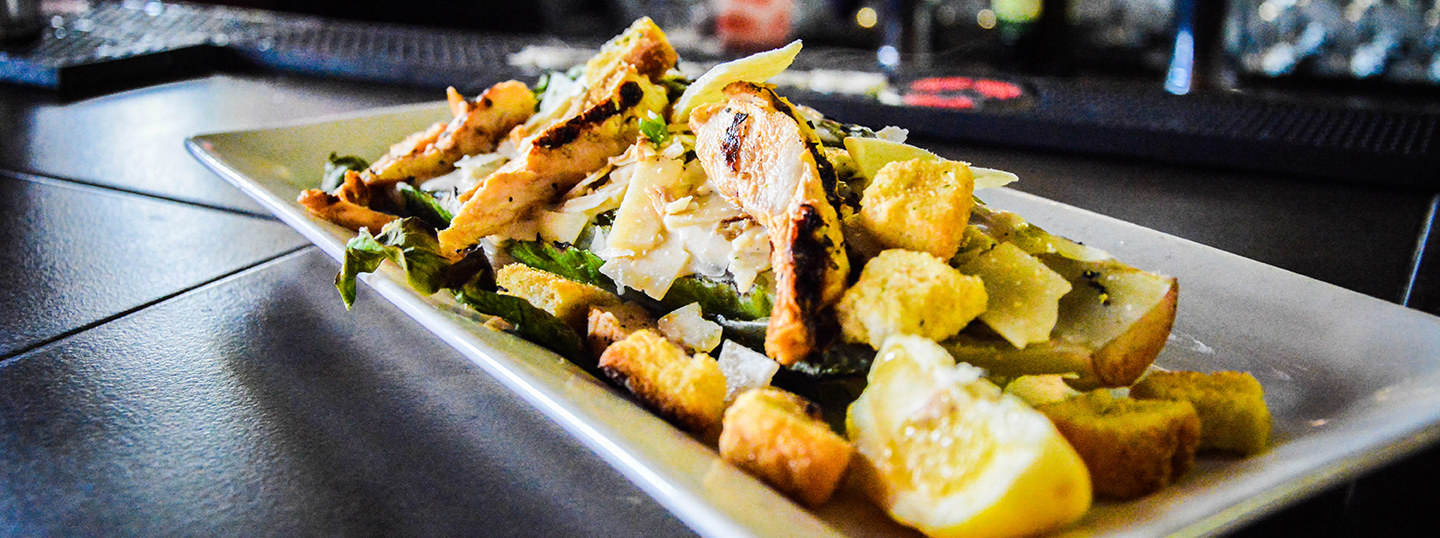 Grilled chicken salad with shaved cheese, croutons, and roasted potatoes on a rectangular white plate at a bar counter