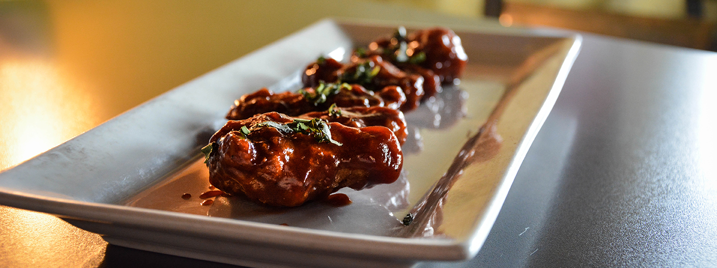 Row of glazed chicken drumsticks in dark barbecue sauce garnished with herbs on a white rectangular plate, rich brown and amber tones